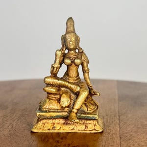 Puede incluir: Pequeña estatua dorada de una figura sentada con detalles ornamentados. La figura está sentada sobre una base detallada. La estatua tiene una apariencia desgastada, lo que sugiere antigüedad. La figura lleva un tocado y joyas. Es probable que sea un objeto religioso o decorativo.