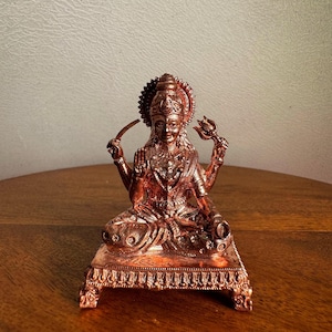 Puede incluir: Una estatua de color cobre de una deidad sentada, adornada con detalles intrincados. La figura sostiene una cuchilla curva en una mano y un tridente en la otra. La estatua está montada sobre una base decorativa, mostrando un tema religioso tradicional.