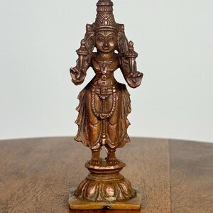 May include: A bronze-colored statue of a deity, featuring intricate details in its clothing and headdress. The figure stands on a detailed pedestal, with arms outstretched. The statue is approximately 5 inches tall, showcasing a traditional design.