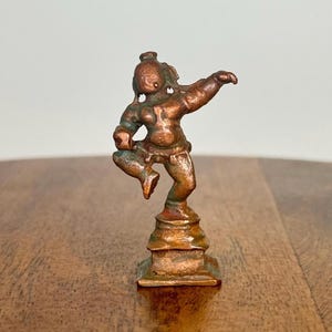 Könnte beinhalten: Kleine Bronzestatue einer tanzenden Figur. Die Figur ist mit einem erhobenen Bein und ausgestreckten Armen posiert. Die Statue hat eine grün-braune Patina und ist auf einem quadratischen Sockel montiert.
