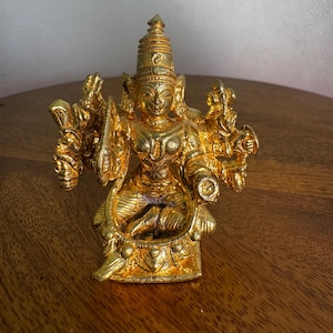 Può includere: Una statua dorata di una divinità, seduta con più braccia e un'acconciatura ornata. La statua è molto dettagliata con intagli intricati e una finitura lucida. La figura è posizionata su una superficie di legno.