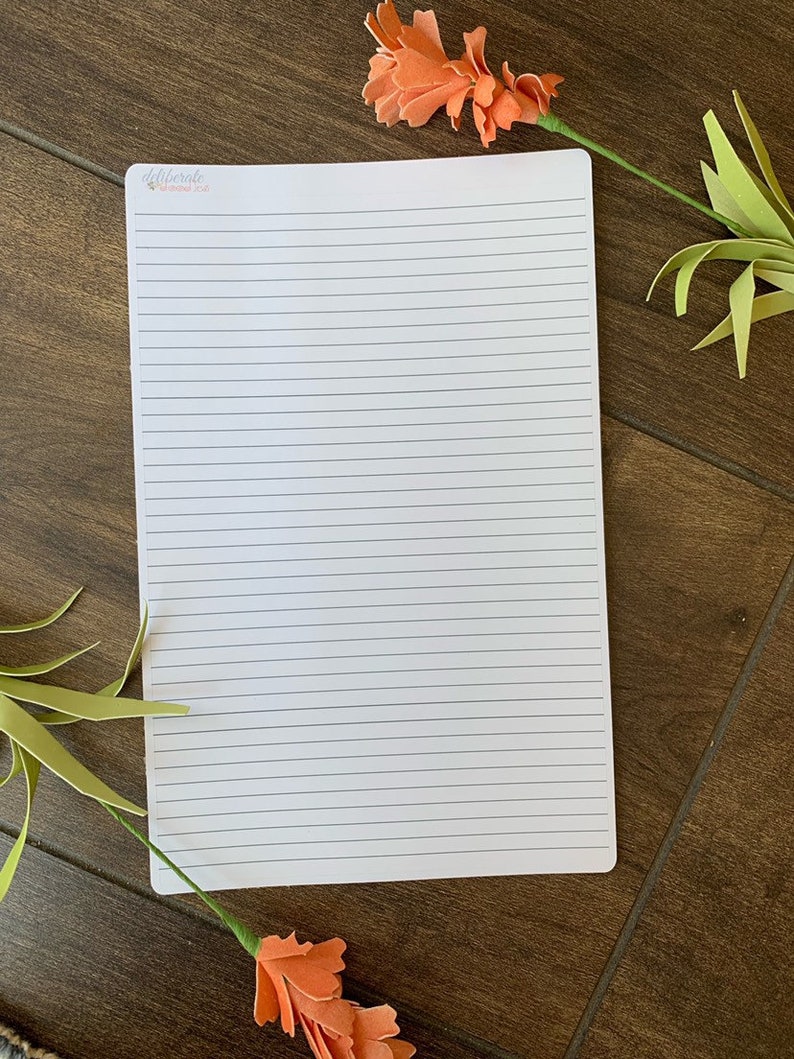 May include: A white lined notebook with the text "deliberate" in pink at the top of the page. The notebook is lying on a brown wooden surface with two orange flowers on either side.