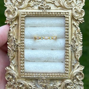 May include: A gold-toned ring with a delicate design, featuring alternating oval-shaped pink stones and small blue stones. The ring is displayed in a gold ornate frame with a white velvet interior, set against a blurred green background.