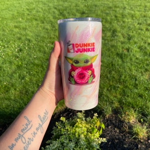 Puede incluir: Un vaso de acero inoxidable blanco y rosa con purpurina, con una imagen de dibujos animados de un personaje verde sosteniendo una rosquilla rosa. El texto "Dunkie Junkie" est&aacute; impreso en el vaso.