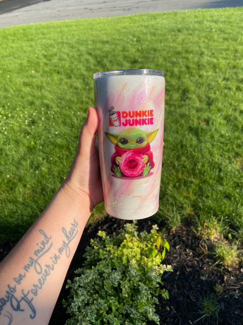 Puede incluir: Un vaso de acero inoxidable blanco y rosa con purpurina, con una imagen de dibujos animados de un personaje verde sosteniendo una rosquilla rosa. El texto "Dunkie Junkie" est&aacute; impreso en el vaso.