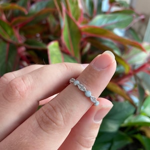 May include: A silver ring with a delicate design featuring small, round, clear stones and oval-shaped, light blue stones. The ring is worn on a finger, with a blurred background of green and red foliage.