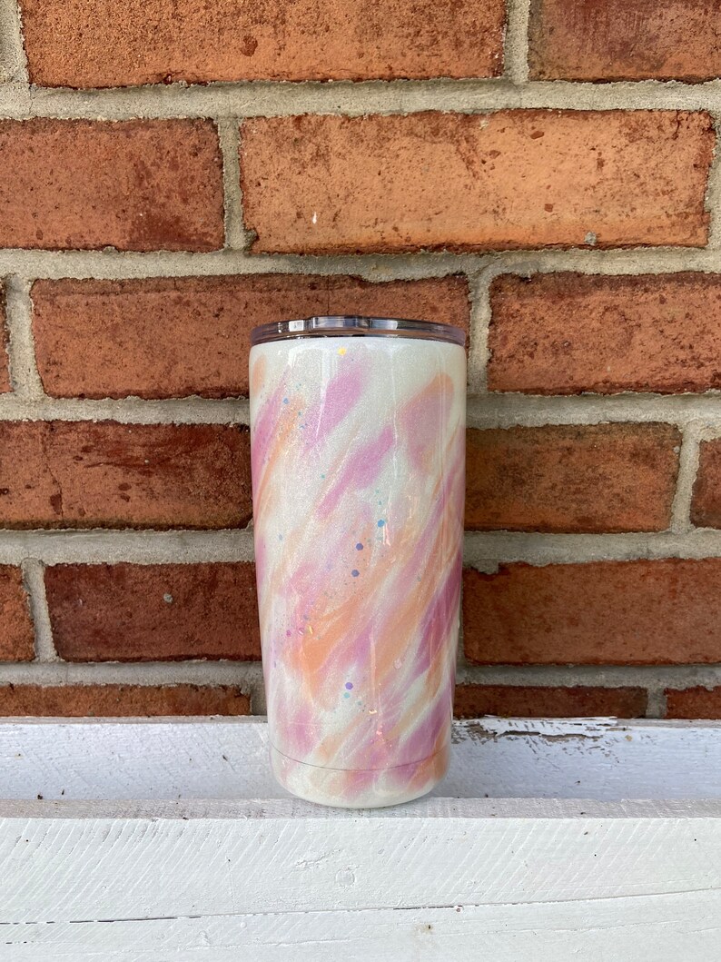 Puede incluir: Un vaso blanco con un dise&ntilde;o de remolino rosa y naranja y detalles de purpurina.