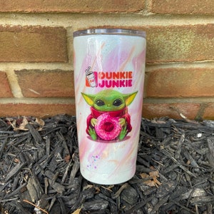 Puede incluir: Un vaso blanco con un dise&ntilde;o de remolino rosa y blanco. El vaso presenta una imagen de dibujos animados de un extraterrestre verde sosteniendo una rosquilla rosa. El texto "Dunkie Junkie" est&aacute; impreso en el vaso.