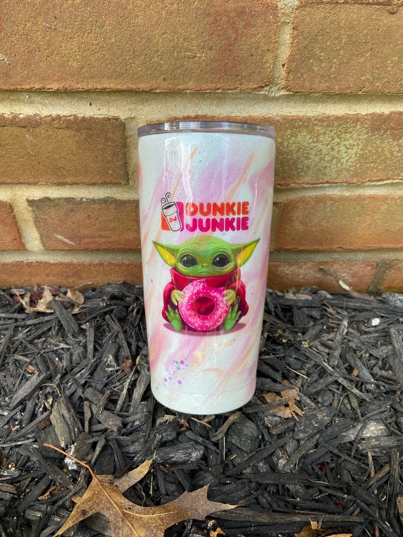 Puede incluir: Un vaso blanco con un dise&ntilde;o de remolino rosa y blanco. El vaso presenta una imagen de dibujos animados de un extraterrestre verde sosteniendo una rosquilla rosa. El texto "Dunkie Junkie" est&aacute; impreso en el vaso.