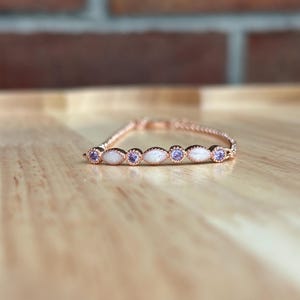 May include: A rose gold ring with a delicate chain design. The ring features alternating oval-shaped white stones and round purple stones. The ring is displayed on a wooden surface.
