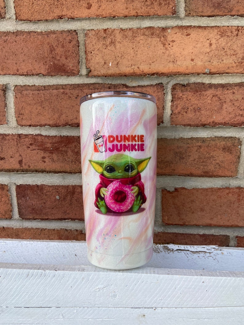 Puede incluir: Un vaso blanco con un dise&ntilde;o de remolino rosa y naranja. El vaso presenta una imagen de dibujos animados de un extraterrestre verde sosteniendo una rosquilla rosa. El texto "Dunkie Junkie" est&aacute; impreso en el vaso.