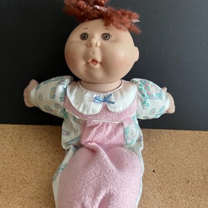 May include: A Cabbage Patch Kid doll with curly red hair wearing a pink and white sleeper with a blue bow. The doll has a round face with rosy cheeks and big brown eyes.