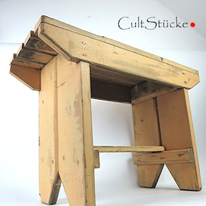 May include: A rustic wooden stool with a light yellow finish. The stool has a simple design with a wide top and two angled legs on each side. The stool is made of reclaimed wood and has a distressed look.