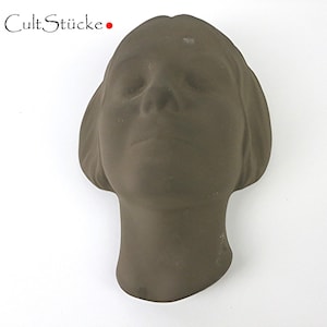 May include: A brown sculpture of a human head with closed eyes and short hair. The sculpture has a matte finish and the text "Cult Stücke" is in the upper left corner.