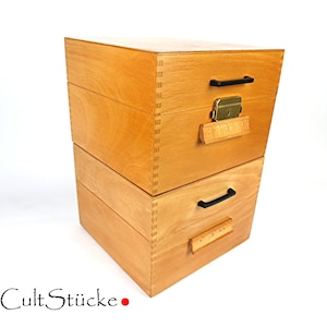 Könnte beinhalten: Ein Stapel aus zwei hellen Holzaufbewahrungsboxen mit sichtbaren Fingerzinken an den Ecken. Jede Box verfügt über einen schwarzen horizontalen Griff und eine kleine Holzlasche. Die obere Box enthält einen goldenen Verschluss. Text CultStücke.