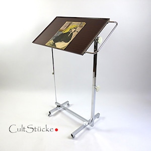 May include: A brown, adjustable display stand with a chrome frame and castors. The stand has a rectangular, angled top with a framed image. The text "Cult Stücke" is visible at the bottom.