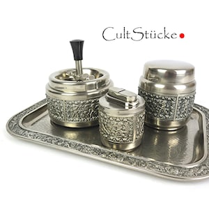 Pode incluir: Um conjunto de fumador prateado vintage, incluindo um cinzeiro, um isqueiro e um recipiente, apresentado numa bandeja retangular. O conjunto apresenta detalhes texturizados e o texto "CultStücke".