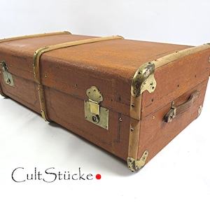May include: Brown vintage trunk with brass hardware, corner protectors, and a leather handle. The trunk has a rectangular shape and is closed with a latch. The text "Cult Stücke" is visible on the bottom left.