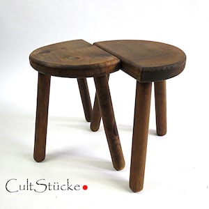 May include: Two connected, half-circle wooden stools with three legs each. The stools are made of dark brown wood and have a rustic, handcrafted appearance. The text "Cult Stücke" is visible at the bottom left.