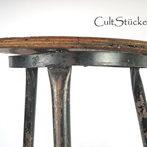 Op de afbeelding: Een vintage kruk met een ronde houten zitting en een zwart metalen frame. De zitting heeft een natuurlijke houtnerf. De metalen poten vertonen tekenen van slijtage. De tekst "CultStücke" is zichtbaar.