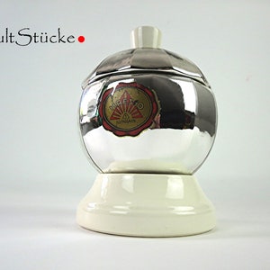 Könnte beinhalten: Eine verchromte Kaffeekanne mit weißem Sockel und Deckel. Die Kanne hat eine reflektierende Oberfläche und ein Vintage-Etikett. Der Text "Cult Stücke" steht über der Kanne.