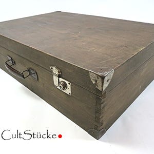 Könnte beinhalten: Ein rechteckiger Holzkoffer in Dunkelbraun mit Metallecken, einem Griff und einem Schnappverschluss. Der Koffer hat Metallbeschläge und der Text "Cult Stücke" ist unten links sichtbar.