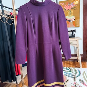 May include: A long-sleeved, knee-length dress in a deep purple hue. The dress features a high, rounded neckline and vertical seams. The hemline is accented with horizontal stripes of gold and peach. The dress is hanging on a wooden hanger.