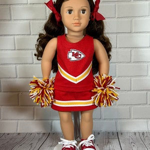 May include: A doll wearing a red and yellow cheerleader outfit with the Kansas City Chiefs logo. The doll has brown hair and is wearing red sneakers.