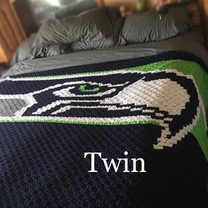 Könnte beinhalten: Eine gehäkelte Decke in Einzelbettgröße mit dem Logo der Seattle Seahawks. Die Decke ist hauptsächlich marineblau, mit dem Logo in Weiß, Grün und Grau. Das Wort "Twin" wird in Weiß angezeigt.