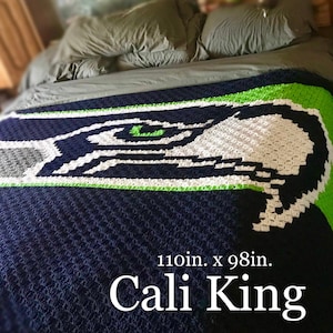 May include: A large crochet blanket featuring a dark blue, white, and neon green graphic of a bird head logo. The blanket measures 110in. x 98in. and is labeled as Cali King. The textured yarn pattern creates a pixelated appearance for the design.