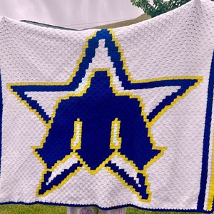 May include: A white crochet blanket with a large blue star design, edged with blue and yellow. The star has yellow and white detailing. The blanket is displayed against a green background.
