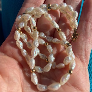 Puede incluir: Un collar de perlas blancas con cuentas doradas. Las perlas son de forma irregular y tienen un ligero brillo iridiscente.