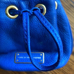 May include: A vibrant cobalt blue leather drawstring bag with gold-coloured hardware. The bag has a drawstring closure, a small rectangular plaque with text, and a top handle. The leather has a textured finish.