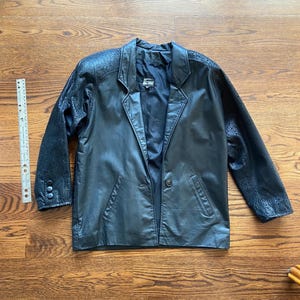 May include: Black leather blazer with patterned sleeves and a single-button closure. The blazer has a notched collar and two front pockets. A ruler is next to the blazer. The blazer is approximately 28 inches long.