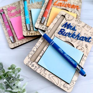 以下が含まれることがあります： ペンと付箋が付いた、木製のパーソナライズされたメモ帳ホルダー。各ホルダーには、カラフルなフォントで名前が記載されています。ホルダーには、ペン、付箋パッド、数学記号のレーザーエッチングデザインがあります。