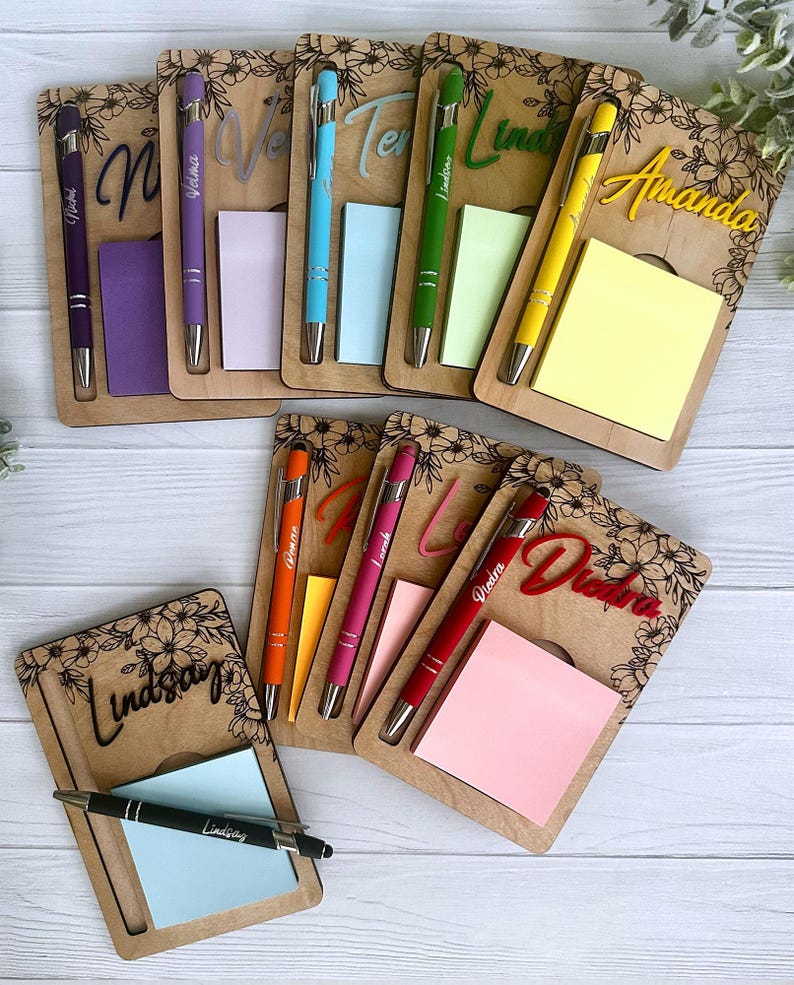 K&ouml;nnte beinhalten: Ein Set personalisierter Notizbl&ouml;cke aus Holz mit Stiften. Jeder Notizblock hat einen anderen Namen und ein Blumenmuster. Die Notizbl&ouml;cke sind in verschiedenen Farben, darunter Lila, Rosa, Gelb, Gr&uuml;n und Blau.