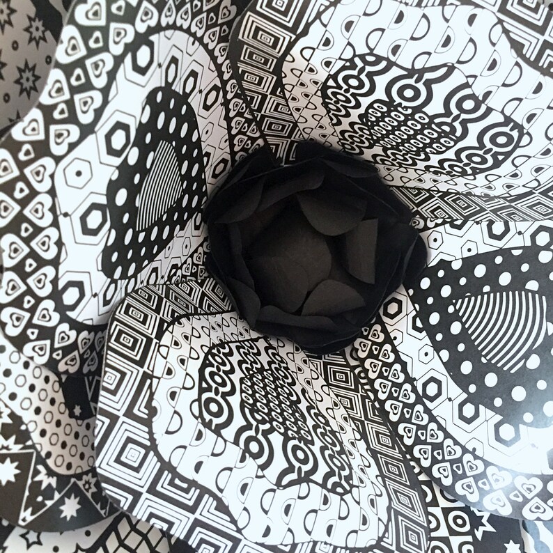 May include: Black and white paper flower with a geometric pattern. The flower is made of multiple layers of paper, each with a different pattern. The center of the flower is a black circle.