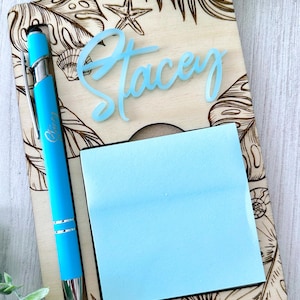 Può includere: Un portablocco in legno con un design a foglie tropicali e conchiglie. È dotato di una penna azzurra e un blocco note adesivo azzurro. Il nome "Stacey" è scritto in corsivo azzurro. La penna ha la parola "Oliver".
