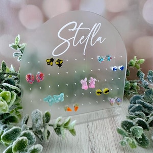 Könnte beinhalten: Transparenter Ohrring-Displayständer aus Acryl mit dem Namen "Stella" in weißer Schrift. Der Ständer hält verschiedene bunte Ohrstecker, darunter Bienen, Hasen und Blumen. Der Ständer steht auf einer Holzoberfläche mit Grün.