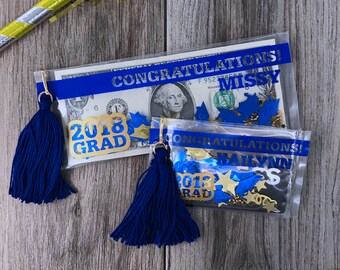 May include: Two clear plastic bags with blue ribbon and gold tassels. The bags contain confetti, a dollar bill, and gold graduation decorations. The bags are labeled "Congratulations Missy" and "Congratulations Bailynn".
