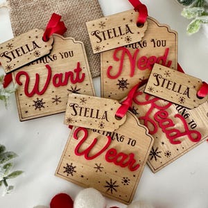 Op de afbeelding: Vier houten cadeau-etiketten met rode letters en sneeuwvlokontwerpen. De etiketten luiden "Stella", "Something You Want", "Something You Need" en "Something To Wear".