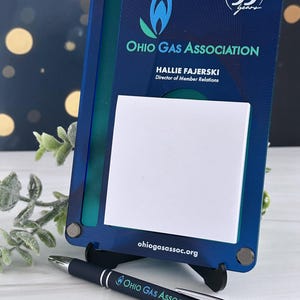 May include: A rectangular blue acrylic desk sign featuring the Ohio Gas Association logo, the text "Celebrating 55 years", "Hallie Fajerski", "Director of Member Relations", and "ohiogasassoc.org", with a white sticky note and a matching blue pen.