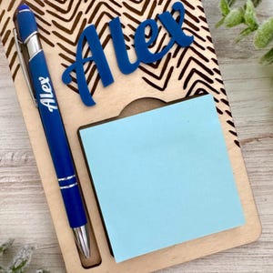 Op de afbeelding: Een houten bureau-organizer met een blauwe pen en een blauwe plakbriefjesblok. De organizer heeft een chevron-patroon en de naam "Alex" is gegraveerd op de voorkant.