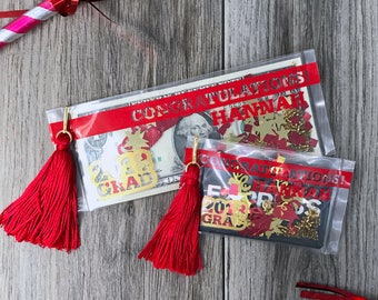 May include: Two clear plastic bags with red tassels, gold confetti, and red and gold paper cutouts. The bags are labeled "Congratulations Hannah!" and "Congratulations! 2023 Grad".