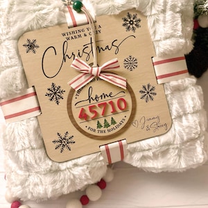 Pode incluir: Uma manta de pele sintética branca com uma placa de madeira que diz "Wishing you a warm & cozy Christmas." A placa tem uma fita vermelha e branca listrada e um ornamento redondo de madeira que diz "There's no place like home 45710 for the holidays." O ornamento tem números vermelhos e um coração vermelho com o texto "Jimmy & Stacey".