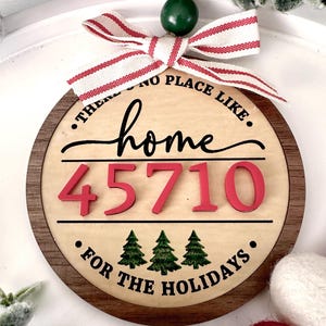 Puede incluir: Adorno navideño redondo de madera con centro de madera clara y borde marrón oscuro. El adorno presenta las palabras "There's no place like home" y "For the Holidays". También tiene el número "45710" en rojo.