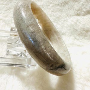 May include: A translucent, light-colored bangle bracelet with a marbled appearance. The bracelet has a smooth, rounded shape and is displayed on a clear acrylic stand. The color palette includes shades of gray, beige, and white.