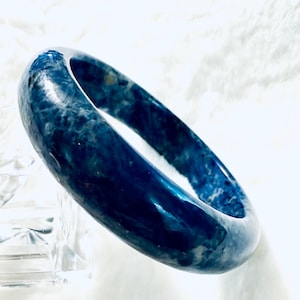 May include: A polished, blue and white marbled stone bangle bracelet. The bracelet is a solid band with a smooth, glossy finish. The color is a deep, rich blue with white veining throughout. The bracelet is displayed on a white surface.