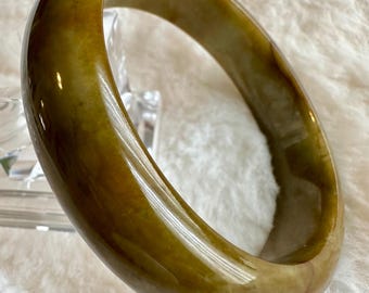 May include: A green jade bangle bracelet with a smooth, polished finish. The bangle has a light brown hue and a slightly oval shape.