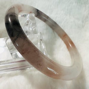 May include: A translucent jade bangle bracelet with a gradient of colors. The bracelet transitions from a dark grey to a light brown and then to a clear, almost white color. The smooth, polished surface reflects light.